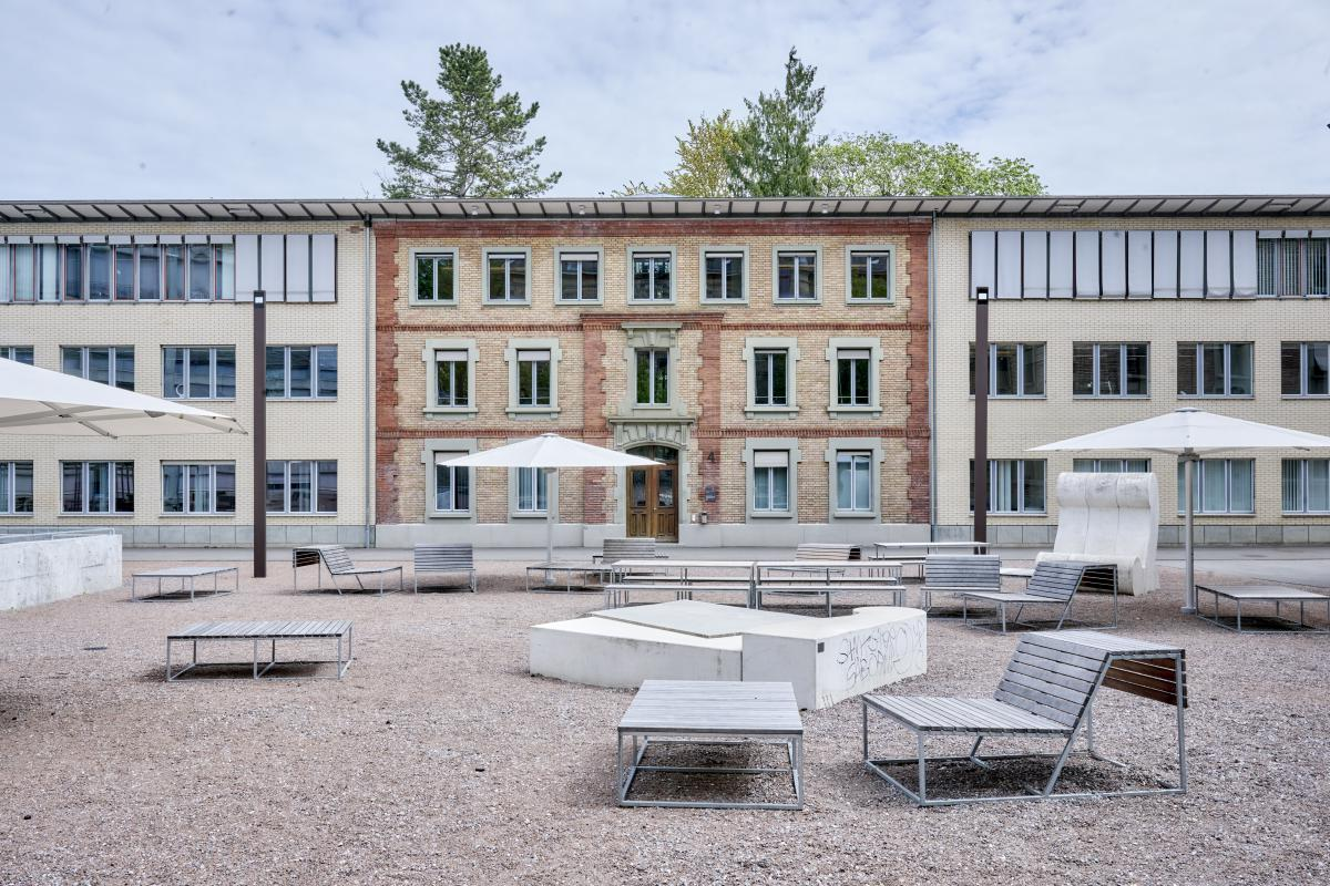 Uni Engehalde – Im Innenhof des Areals Tische und Bänke, aufgespannter Sonnenschirm. Gebäude im Hintergrund