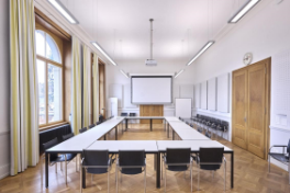 H6 – Sitzungszimmer 101 – Tische im Rechteck aufgestellt mit schwarzen Ledersesseln – im Vordergrund hängt Leinwand herunter, Beamer oben an Decke, links Fensterfront, rechts Eingangstüre.