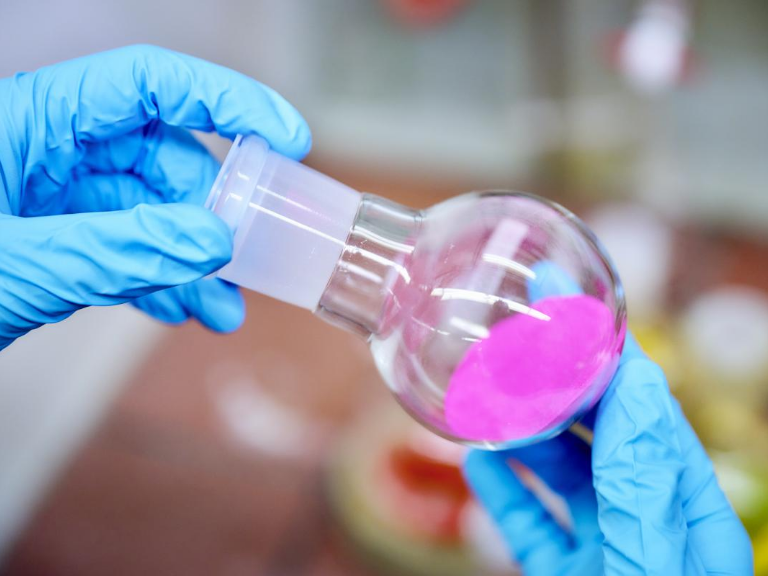 Hand mit blauem Handschuh hält einen Glasbehälter mit pink-farbiger Substanz